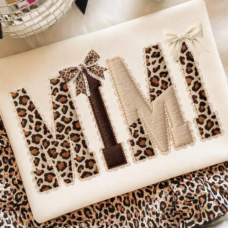 May include: Cream-colored decorative pillow with the word "MIMI" embroidered in a mix of leopard print, brown, and beige fabrics. Each letter is outlined with a delicate lace trim, and small bows adorn the "M" and "I".