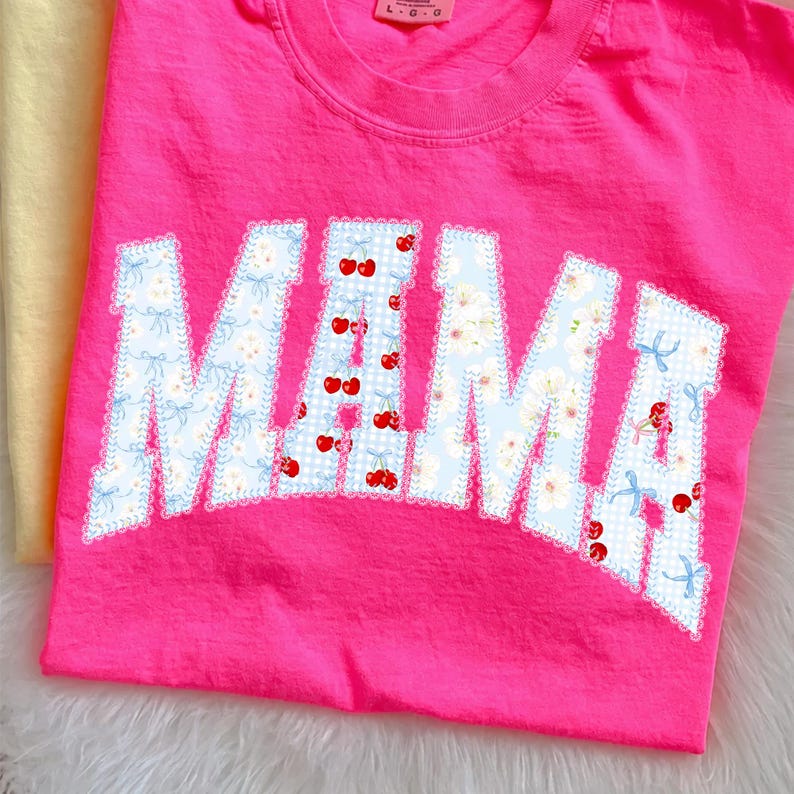 May include: A bright pink t-shirt with the word "MAMA" in a white, blue, and red floral design. The letters are outlined with a white lace trim. The shirt is made of a soft material.
