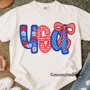 May include: White t-shirt with a patriotic design. The design features the letters "USA" in red, white, and blue. The "U" is blue with floral accents, the "S" is red gingham, and the "A" is blue with white polka dots and a red bow.