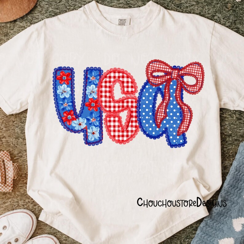 May include: White t-shirt with a patriotic design. The design features the letters "USA" in red, white, and blue. The "U" is blue with floral accents, the "S" is red gingham, and the "A" is blue with white polka dots and a red bow.