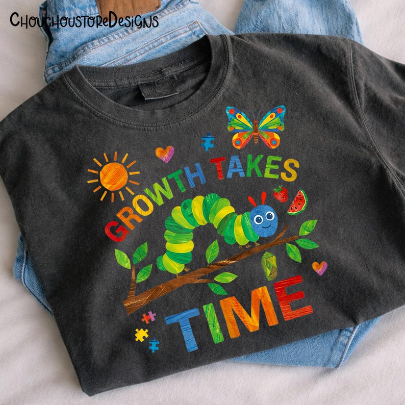 Puede incluir: Camiseta gris oscuro con un gr&aacute;fico colorido de una oruga, una mariposa, un sol y las palabras "GROWTH TAKES TIME". El dise&ntilde;o incluye una rodaja de sand&iacute;a y corazones. La camiseta se muestra sobre unos vaqueros azules.
