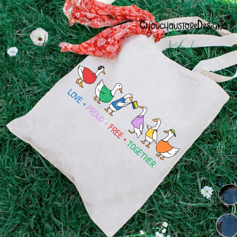 May include: A cream-colored tote bag with a colorful graphic of ducks in various outfits, including a rainbow flag. The text on the bag reads "LOVE &bull; PROUD &bull; FREE &bull; TOGETHER". The bag has long, cream-colored handles.