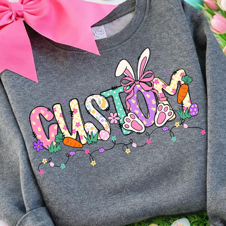 Peut inclure: Un sweat-shirt gris avec le mot "CUSTOM" en lettres color&eacute;es sur le th&egrave;me de P&acirc;ques. Le motif comprend des oreilles de lapin, un n&oelig;ud, des carottes et des &oelig;ufs de P&acirc;ques. Un grand n&oelig;ud rose est visible en haut &agrave; gauche.