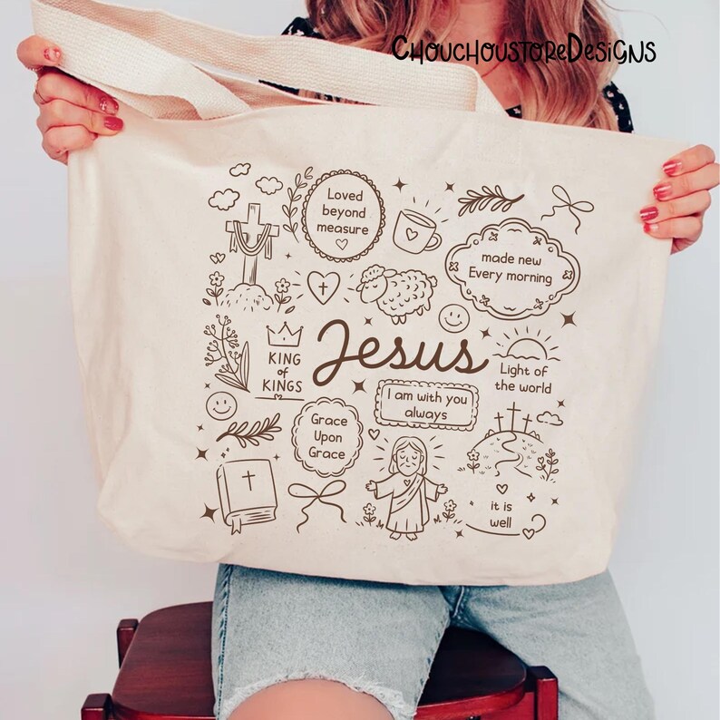 May include: Cream-colored tote bag with a beige handle, featuring a black line art design. The design includes the word "Jesus" and various religious symbols and phrases such as "Loved beyond measure" and "Light of the world".