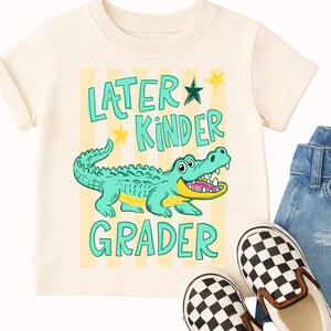 May include: Cream-coloured t-shirt with the words "Later Kinder Grader" and a cartoon alligator. The alligator is teal with a yellow belly and open mouth. Yellow stars and stripes are behind the text and alligator.