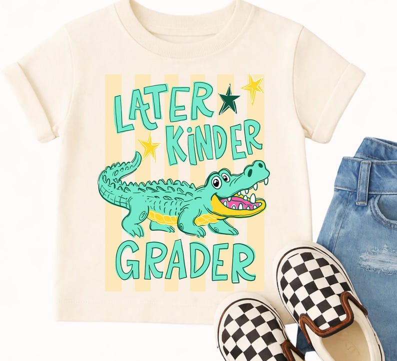 May include: Cream-coloured t-shirt with the words "Later Kinder Grader" and a cartoon alligator. The alligator is teal with a yellow belly and open mouth. Yellow stars and stripes are behind the text and alligator.