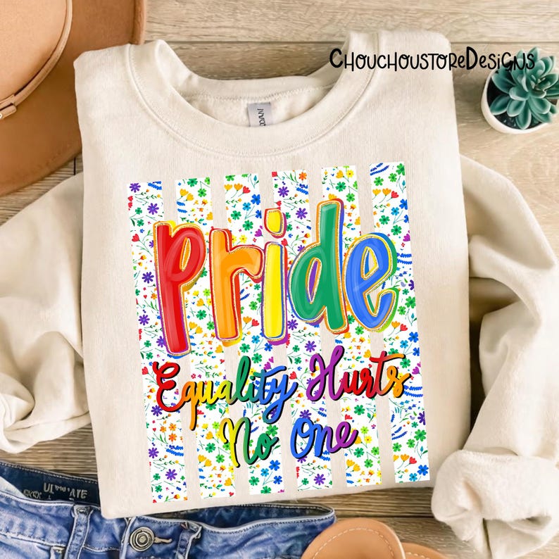 May include: Cream-colored sweatshirt with the word "Pride" in rainbow colors, and the phrase "Equality Hurts No One." The design is set against a floral and striped background. The sweatshirt is folded.