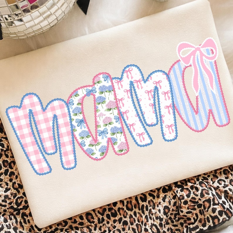 May include: A beige sweatshirt with the word "mama" in a patchwork design. The letters feature pink gingham, floral patterns, and blue and pink striped fabric, all outlined in blue and pink scalloped trim.