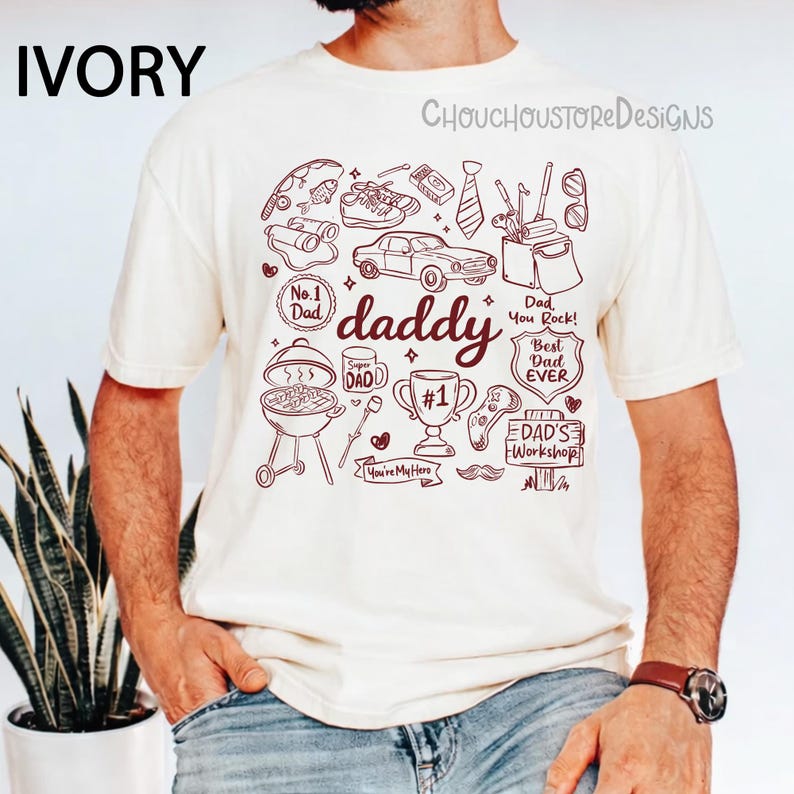 May include: Ivory-colored t-shirt with a maroon graphic featuring the word "daddy" and various illustrations like a car, grill, and trophy. Text includes "No. 1 Dad", "Best Dad Ever", and "Dad, You Rock!"