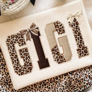 May include: A cream-colored decorative pillow with the name "GIGI" embroidered in a mix of leopard print and brown fabric. The letters are outlined with lace and adorned with small bows. The pillow is set against a soft, white background.