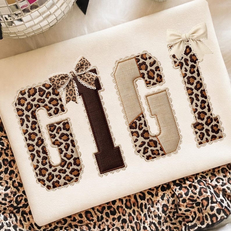 May include: A cream-colored decorative pillow with the name "GIGI" embroidered in a mix of leopard print and brown fabric. The letters are outlined with lace and adorned with small bows. The pillow is set against a soft, white background.