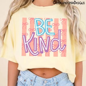 以下が含まれることがあります： 明るい黄色のTシャツ。カラフルなデザインで「BE KIND」の文字がプリントされています。「BE」の文字は白の縁取りのある青色、「KIND」の文字は白の縁取りのある紫色です。背景には、リボンの模様が入ったピンク色のストライプがあります。