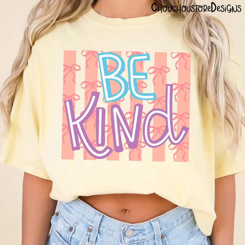 以下が含まれることがあります： 明るい黄色のTシャツ。カラフルなデザインで「BE KIND」の文字がプリントされています。「BE」の文字は白の縁取りのある青色、「KIND」の文字は白の縁取りのある紫色です。背景には、リボンの模様が入ったピンク色のストライプがあります。