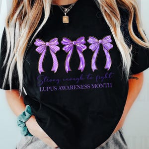 K&ouml;nnte beinhalten: Schwarzes T-Shirt mit drei lila Schleifen mit unterschiedlichen Mustern und metallischen Akzenten. Der Text "Strong enough to fight" und "LUPUS AWARENESS MONTH" ist unter den Schleifen in lila Schrift gedruckt. Ein l&auml;ssiges Kleidungsst&uuml;ck.