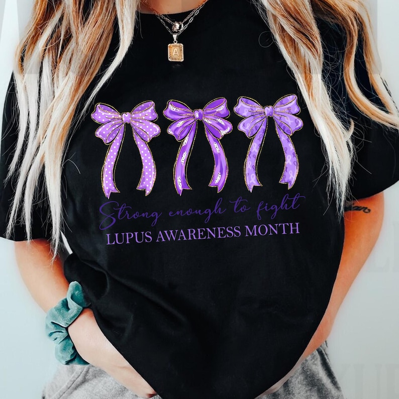K&ouml;nnte beinhalten: Schwarzes T-Shirt mit drei lila Schleifen mit unterschiedlichen Mustern und metallischen Akzenten. Der Text "Strong enough to fight" und "LUPUS AWARENESS MONTH" ist unter den Schleifen in lila Schrift gedruckt. Ein l&auml;ssiges Kleidungsst&uuml;ck.