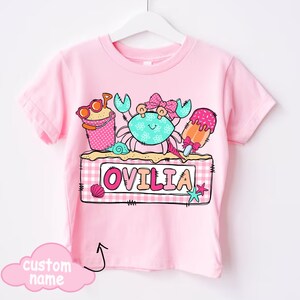 May include: Pink t-shirt featuring a colorful beach-themed graphic. The design includes a crab, ice cream, and a sand pail with sunglasses. The name "OVILIA" is displayed on a sign. A cloud graphic indicates a custom name can be added.