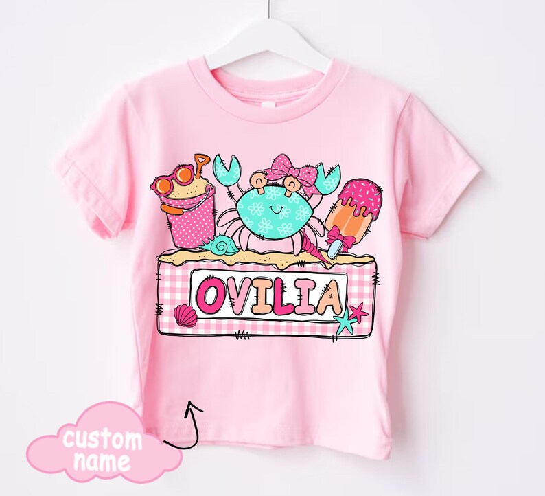 May include: Pink t-shirt featuring a colorful beach-themed graphic. The design includes a crab, ice cream, and a sand pail with sunglasses. The name "OVILIA" is displayed on a sign. A cloud graphic indicates a custom name can be added.