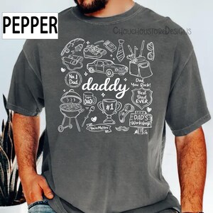 May include: A gray t-shirt with a white graphic design featuring the word "daddy" and various illustrations like a car, trophy, and barbecue. The shirt also includes the text "No. 1 Dad", "Best Dad Ever", and "Dad's Workshop".
