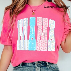 May include: A pink t-shirt with the word "MAMA" in large, decorative letters. The letters have various patterns: blue stripes, blue polka dots, pink floral, and blue floral. The shirt is a vibrant pink color.