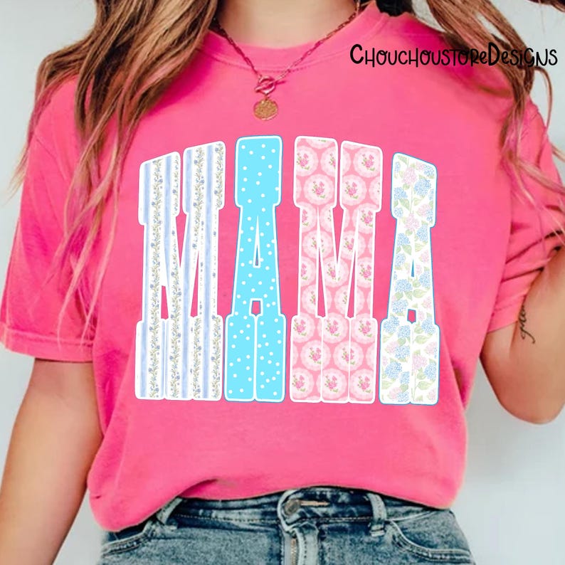 May include: A pink t-shirt with the word "MAMA" in large, decorative letters. The letters have various patterns: blue stripes, blue polka dots, pink floral, and blue floral. The shirt is a vibrant pink color.