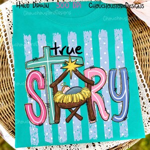 May include: A teal t-shirt featuring a graphic with the text true STORY. The design includes a light blue cross, a yellow star, and a manger scene with a baby in a yellow bed. The background has light blue vertical stripes with white dots.
