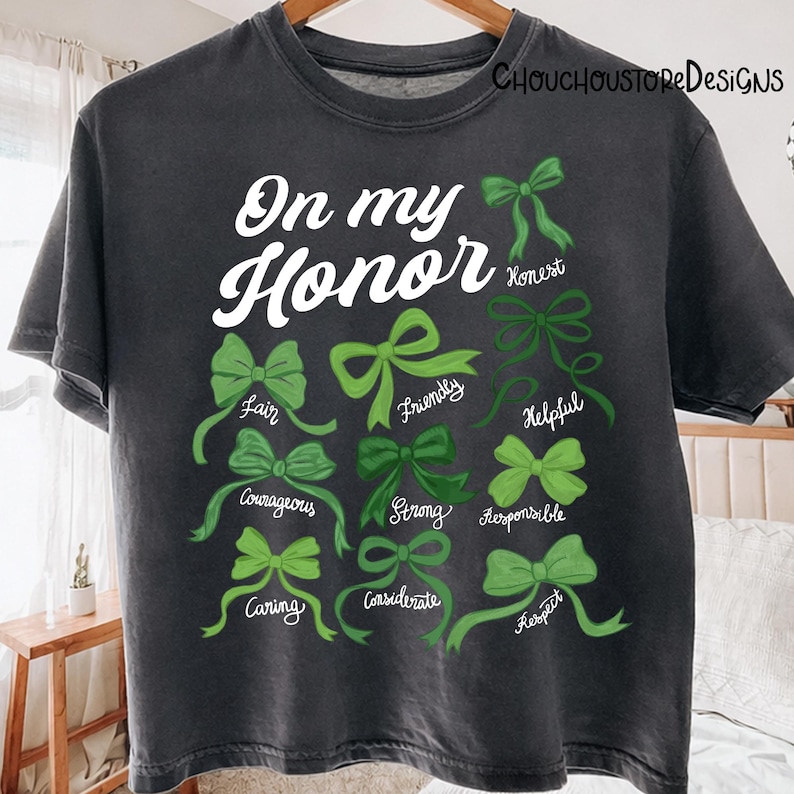 May include: Dark gray t-shirt with the words "On my Honor" in white script. Below, green bows are paired with words like "Fair," "Friendly," and "Courageous." The shirt has a vintage, worn look.