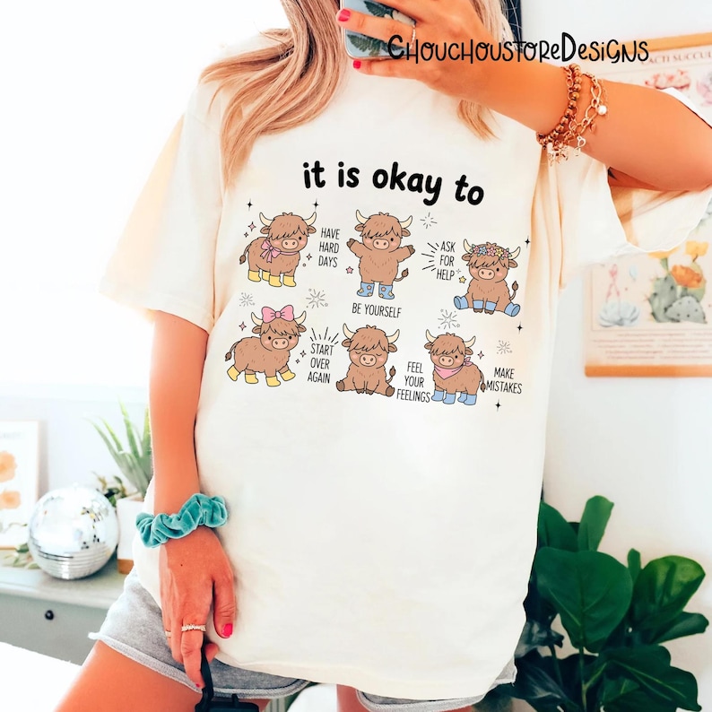 May include: Cream-colored t-shirt with the text "it is okay to" and illustrations of cartoon Highland cows. Each cow has a different accessory and a motivational phrase, such as "Have hard days" and "Ask for help."