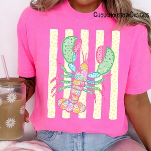 May include: A bright pink t-shirt featuring a colorful lobster design. The lobster is decorated with seashells and other marine elements, set against a backdrop of vertical white and yellow stripes. The text "ChouchouStoreDesigns" is printed on the top right.