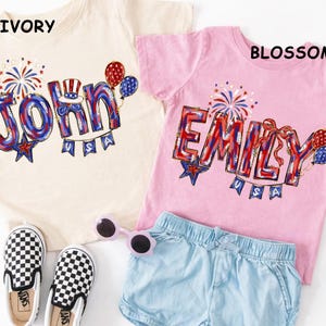 Puede incluir: Dos camisetas infantiles, una marfil y otra rosa, con nombres personalizados en rojo, blanco y azul. La camiseta marfil dice "John" y la rosa "Emily". Ambas camisetas tienen gr&aacute;ficos festivos y la palabra "USA".