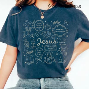 May include: Navy blue t-shirt with a beige graphic design featuring the word "Jesus" and various religious symbols and phrases. The design includes a cross, a heart, a lamb, and the phrase "Light of the world".
