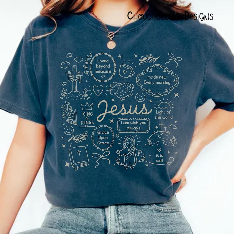 May include: Navy blue t-shirt with a beige graphic design featuring the word "Jesus" and various religious symbols and phrases. The design includes a cross, a heart, a lamb, and the phrase "Light of the world".