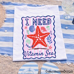 May include: White t-shirt with a colourful graphic design. The design features a red starfish, seashells, and the text "I NEED Vitamin Sea" in a blue and pink frame. The shirt is displayed on a striped beach towel with flip-flops.