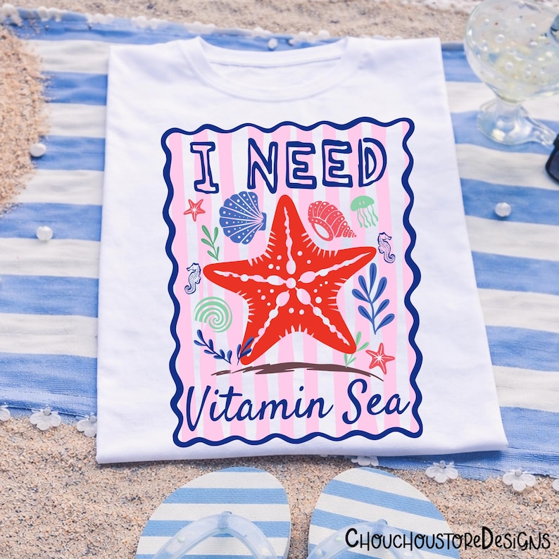 May include: White t-shirt with a colourful graphic design. The design features a red starfish, seashells, and the text "I NEED Vitamin Sea" in a blue and pink frame. The shirt is displayed on a striped beach towel with flip-flops.