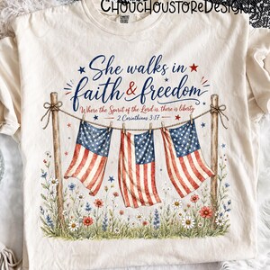 May include: Cream-colored t-shirt featuring the text "She walks in faith & freedom" with American flags and floral accents. The design includes the Bible verse 2 Corinthians 3:17. A patriotic and inspirational graphic tee.
