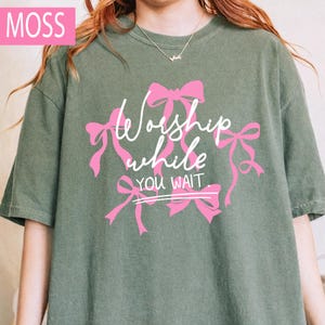 May include: A moss green t-shirt with the words "Worship while you wait" in white script, accented by pink bows. The shirt has a relaxed fit and short sleeves, perfect for casual wear.