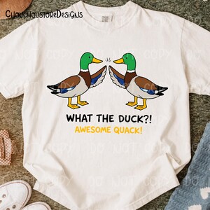 May include: White t-shirt featuring a cartoon design of two ducks facing each other. The ducks have green heads, yellow beaks, and brown and blue wings. The text "WHAT THE DUCK?! AWESOME QUACK!" is below the ducks.