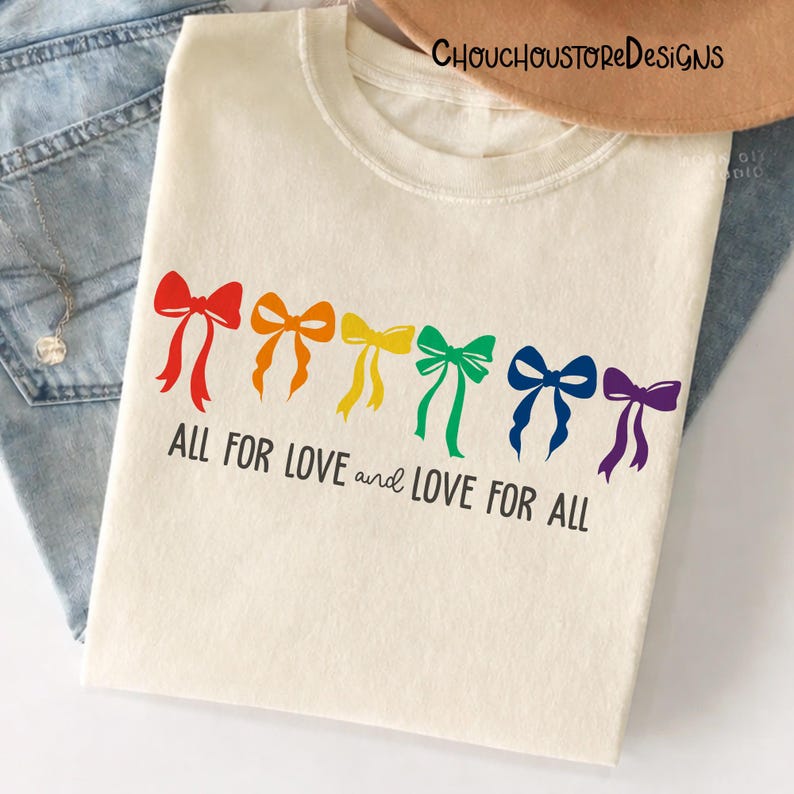 May include: Cream-colored t-shirt with a rainbow of bows in red, orange, yellow, green, blue, and purple. The text "ALL FOR LOVE and LOVE FOR ALL" is printed below the bows. The shirt is displayed on a light background.