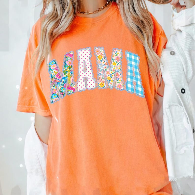 May include: Orange short-sleeved t-shirt with the word "MAMA" in a decorative, arched design. The letters are filled with floral patterns, polka dots, and blue gingham. The shirt is a solid orange colour.