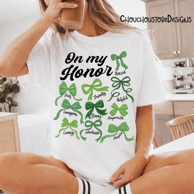 May include: White t-shirt with the text "On my Honor" in black script. Below the text are green bows with words like "Honest", "Fair", and "Courageous". The shirt is worn by a person holding a beverage.
