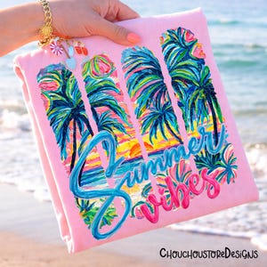 May include: A pink beach towel with a colorful tropical design featuring palm trees and a sunset. The words "Summer Vibes" are written in blue and pink script. The towel is held up near the ocean.