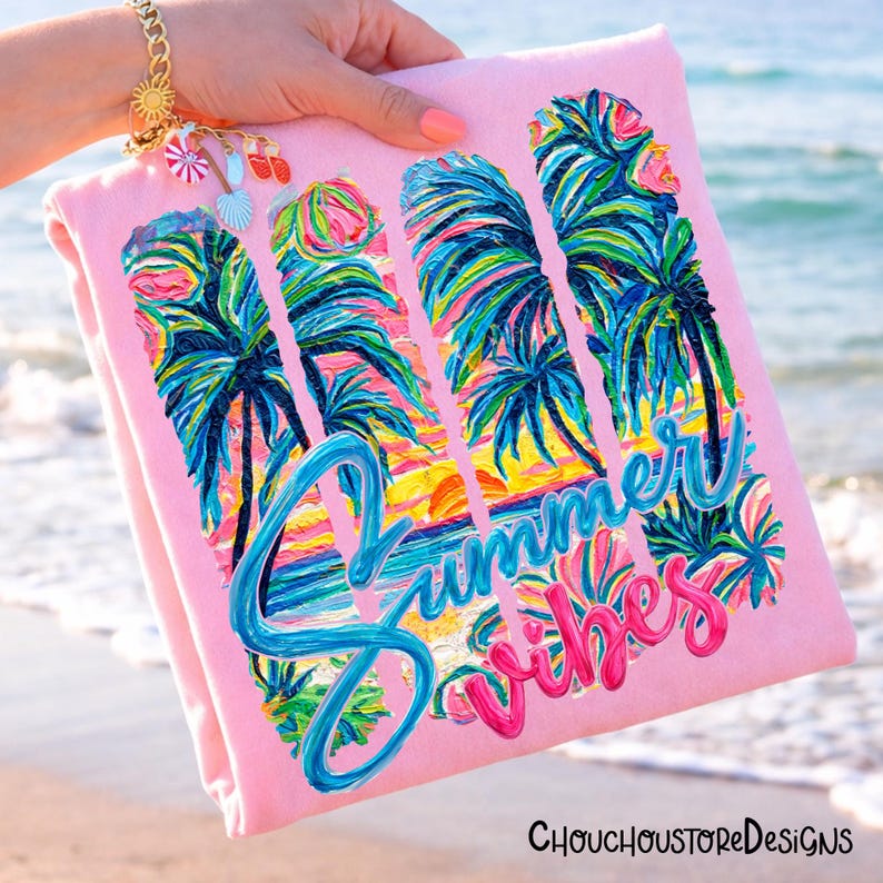 May include: A pink beach towel with a colorful tropical design featuring palm trees and a sunset. The words "Summer Vibes" are written in blue and pink script. The towel is held up near the ocean.