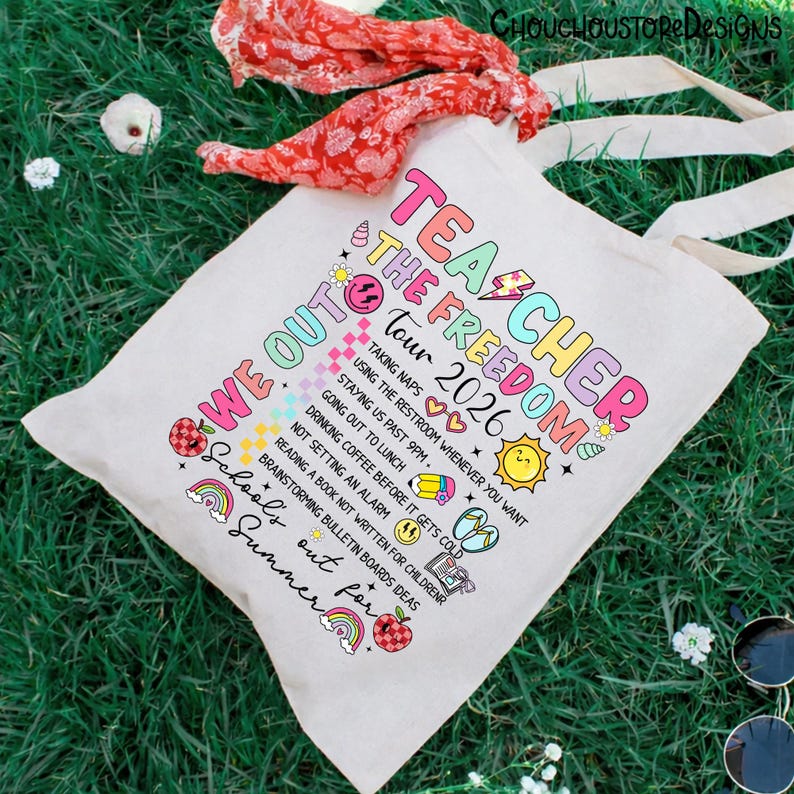 May include: A white canvas tote bag with the text "Teacher The Freedom Tour 2026" in colorful letters. The bag features a list of fun summer activities. A red scarf and sunglasses are on the green grass.