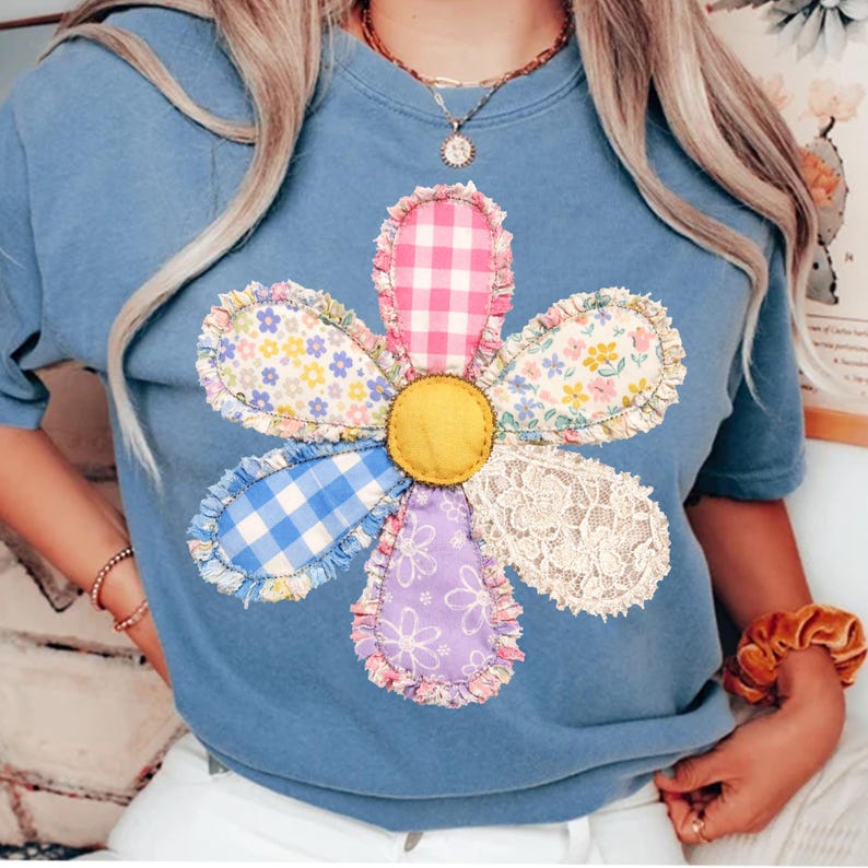 May include: A light blue t-shirt featuring a large, colorful flower applique. The flower has petals of various patterns, including gingham, floral prints, and lace, with a yellow felt center. The shirt is a casual, comfortable style.
