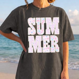 May include: Dark gray t-shirt with the word "SUMMER" in large, white, block letters. The letters are filled with a pastel pattern of pink, blue, and yellow shapes. The background is a beach scene with the ocean and sky.