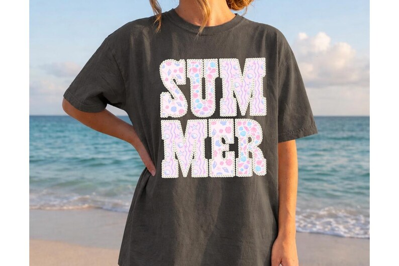 May include: Dark gray t-shirt with the word "SUMMER" in large, white, block letters. The letters are filled with a pastel pattern of pink, blue, and yellow shapes. The background is a beach scene with the ocean and sky.