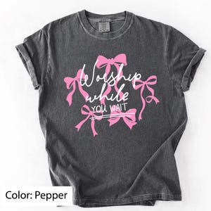 May include: Pepper-colored t-shirt with the text "Worship while you wait" in white script and pink bows. The short-sleeved shirt has a crew neck and a slightly faded appearance.