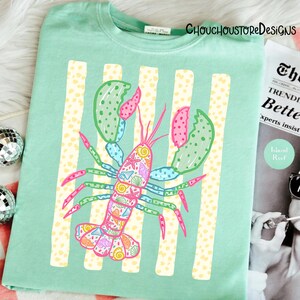 May include: A light green t-shirt featuring a colorful lobster design. The lobster is decorated with seashells and other marine life. Vertical yellow and white stripes are behind the lobster. The text "CHOUCHOUSTOREDESIGNS" is at the top.