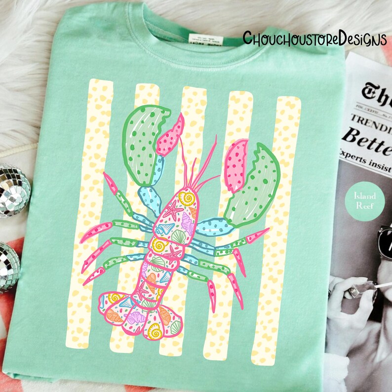 May include: A light green t-shirt featuring a colorful lobster design. The lobster is decorated with seashells and other marine life. Vertical yellow and white stripes are behind the lobster. The text "CHOUCHOUSTOREDESIGNS" is at the top.
