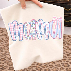 May include: Cream-colored t-shirt with the word "MAMA" in a patchwork design. The letters feature pink gingham, floral, bow, and striped patterns, outlined in blue. A hand is holding the shirt.