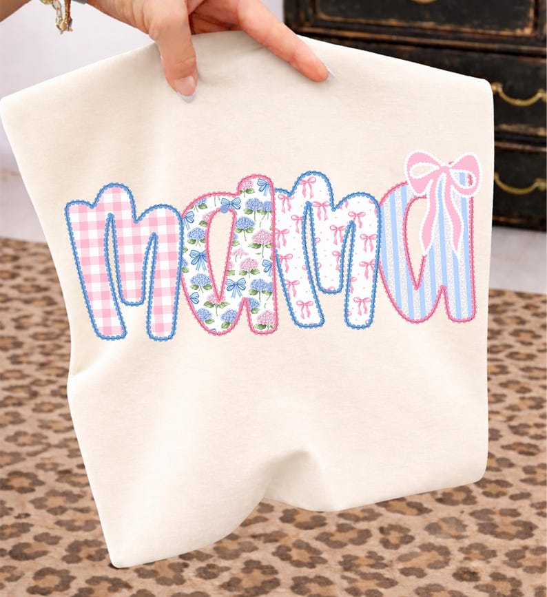 May include: Cream-colored t-shirt with the word "MAMA" in a patchwork design. The letters feature pink gingham, floral, bow, and striped patterns, outlined in blue. A hand is holding the shirt.